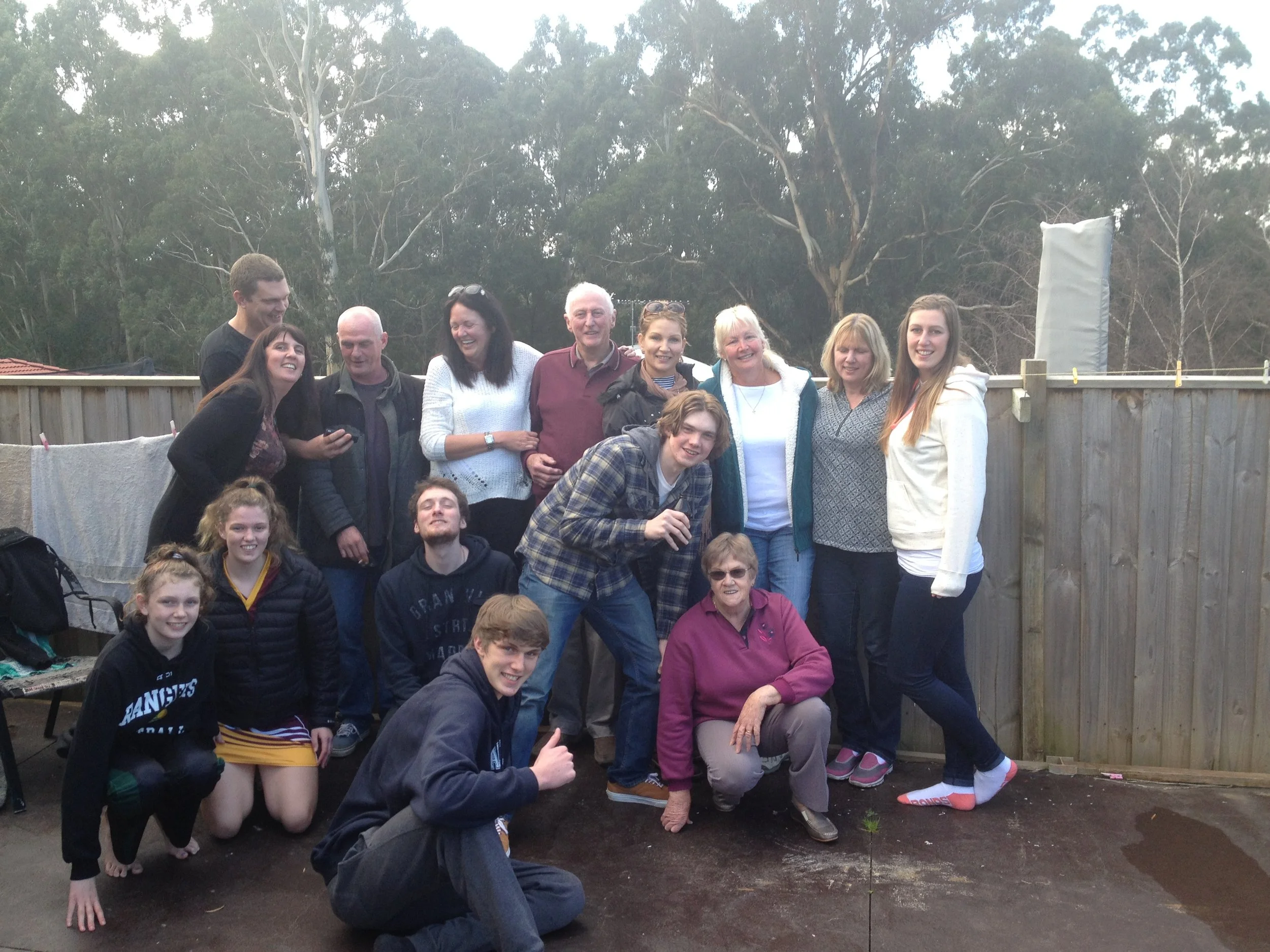 Group of 16 people, including children, teenagers, and adults, gathered outdoors for a group photo, standing or kneeling on dirt ground with a wooden fence and trees in the background.