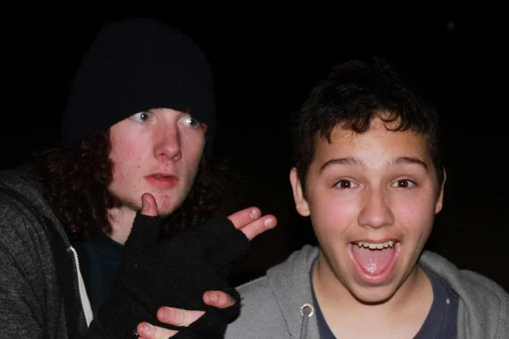 Two young boys outdoors at night, one making a peace sign and the other smiling with tongue out.