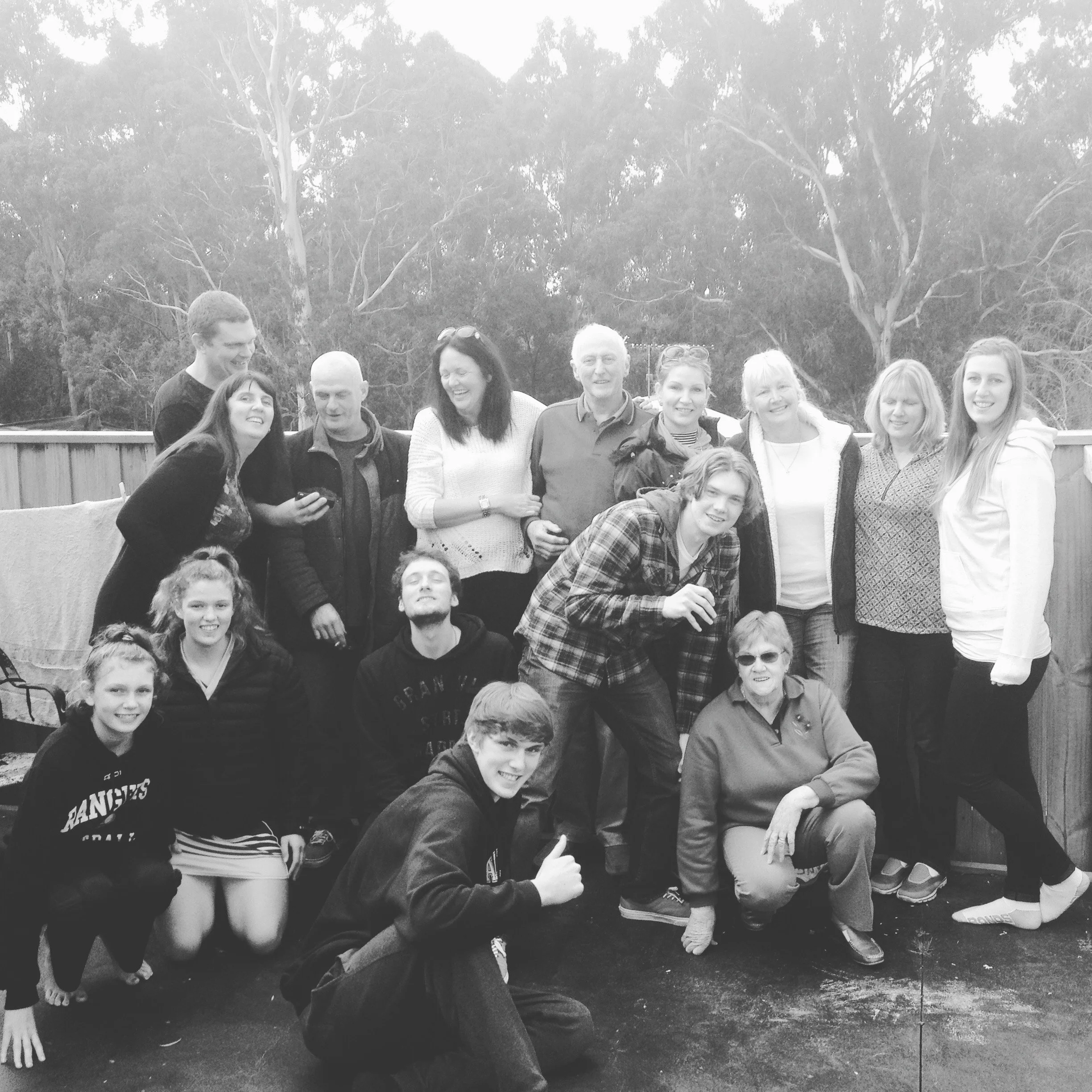 Group of fifteen people, some standing and some kneeling, outdoors on a paved surface with trees and a wooden fence in the background, smiling and posing for a photo.