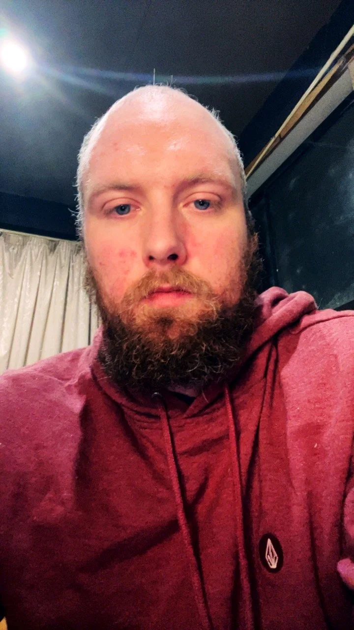 A close-up selfie of a man with short, light-colored hair and a full beard, wearing a red hoodie, indoors at night.