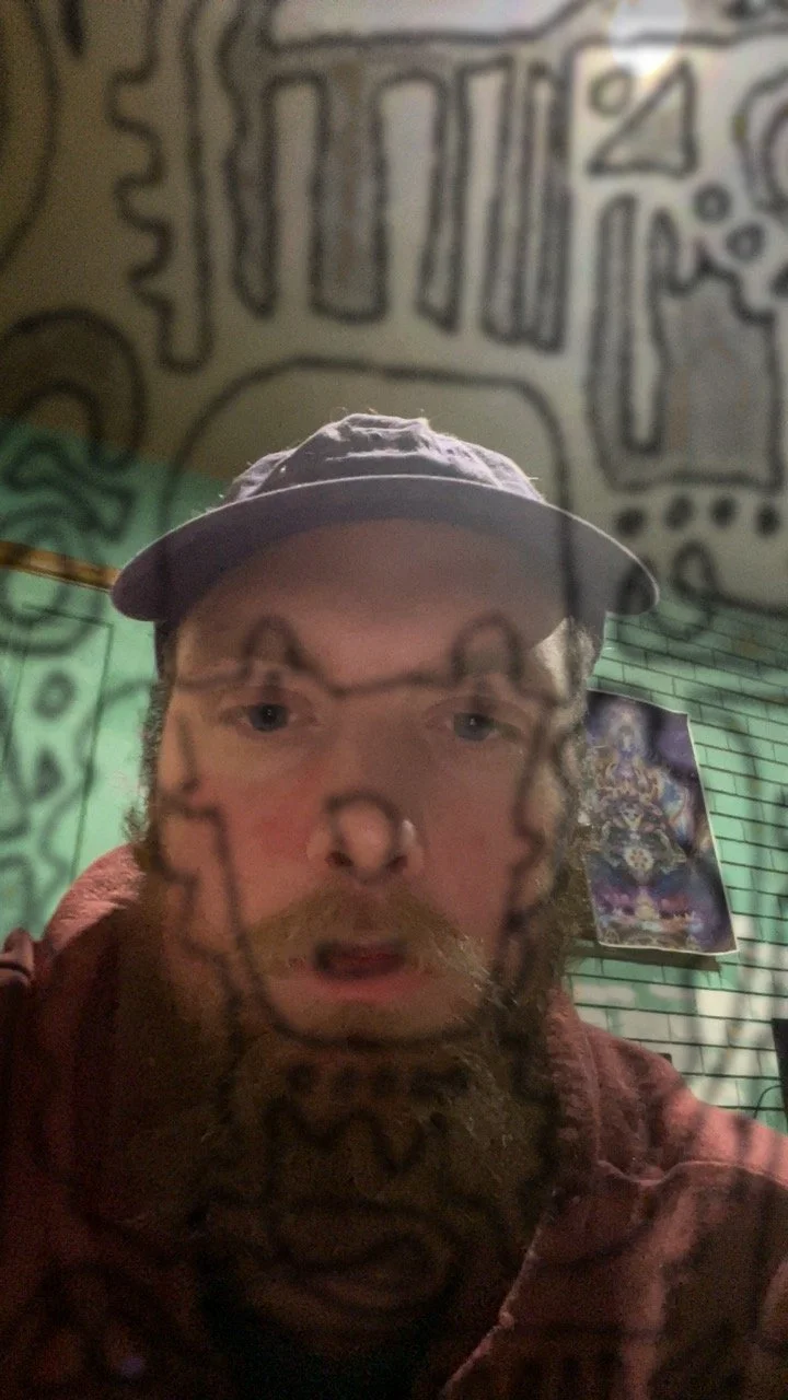 A man with a beard is holding a transparent mask with black outline drawings of a face in front of his face. The background features a green wall with black graffiti-style art and a colorful poster.