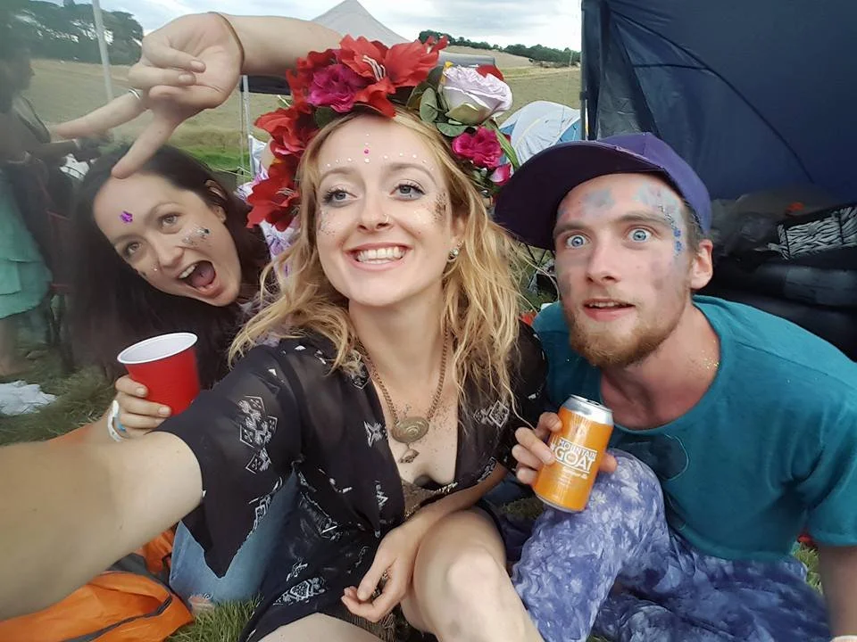 Three friends at an outdoor festival, smiling for a selfie, with colorful face paint, glitter, and a flower crown, surrounded by tents and grass.