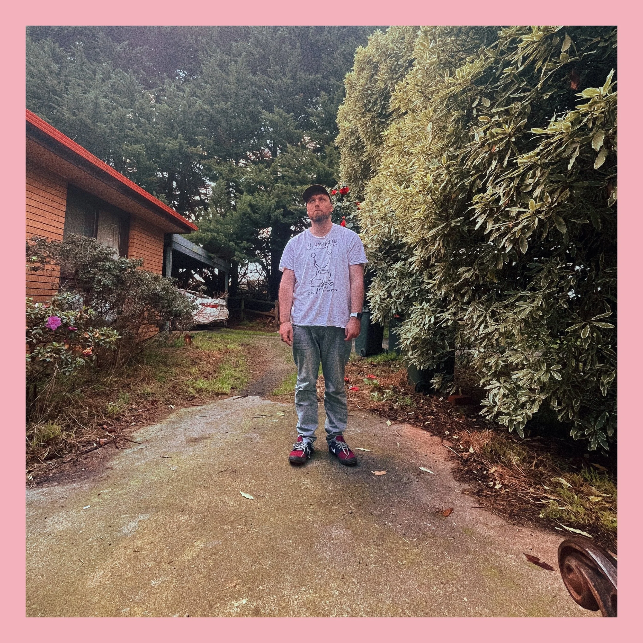A man standing on a paved driveway in a neighborhood with trees and bushes around him, wearing a light gray T-shirt, gray pants, and sneakers, with a dark cap on his head.