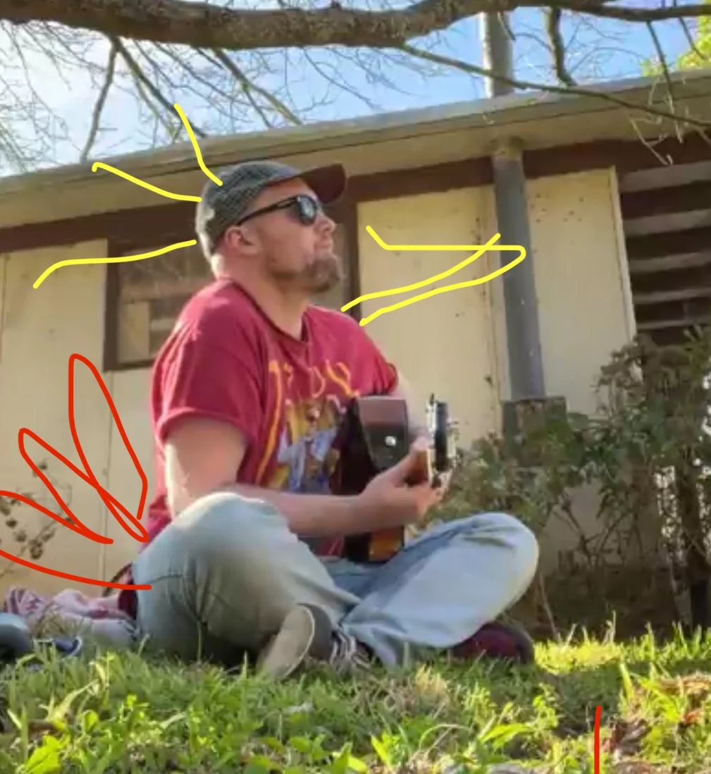 A man sitting cross-legged on grass, wearing a red t-shirt, beige pants, sunglasses, and a hat, playing an acoustic guitar outdoors near a house with a wooden porch, with trees and bushes in the background.