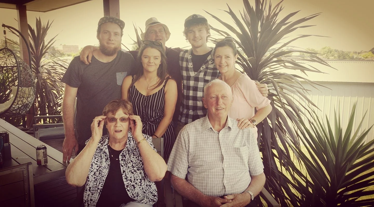 Group of seven people gathered outdoors on a patio, posing for a photo with large plants and a building in the background. Four people are standing in the back row, and three are sitting in front. The group includes a mix of young adults and seniors.