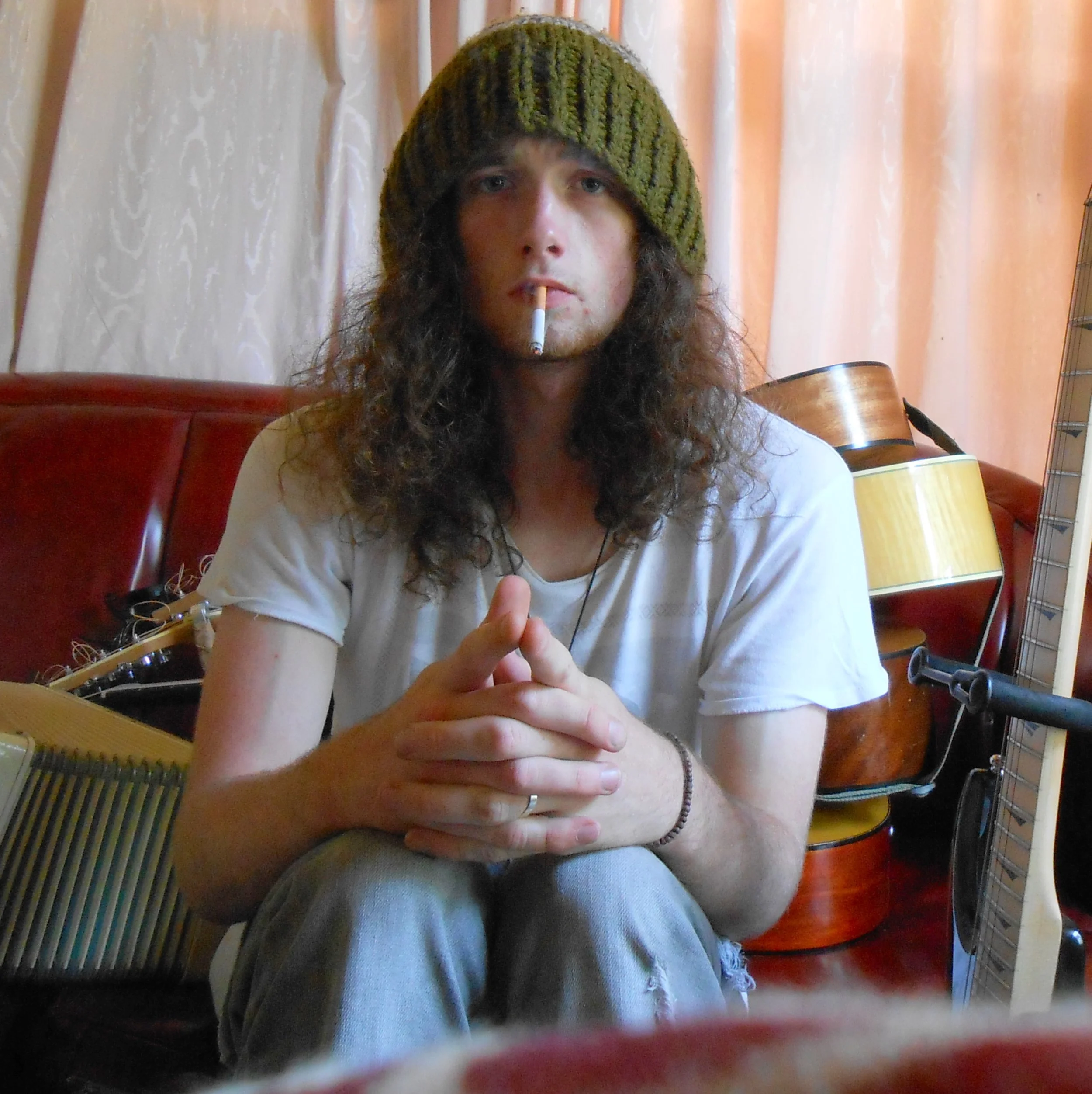 Young man with long curly hair wearing a knit beanie, white t-shirt, and a bracelet, sitting inside with crossed hands, cigarette in his mouth, surrounded by musical instruments including a guitar and a microphone.