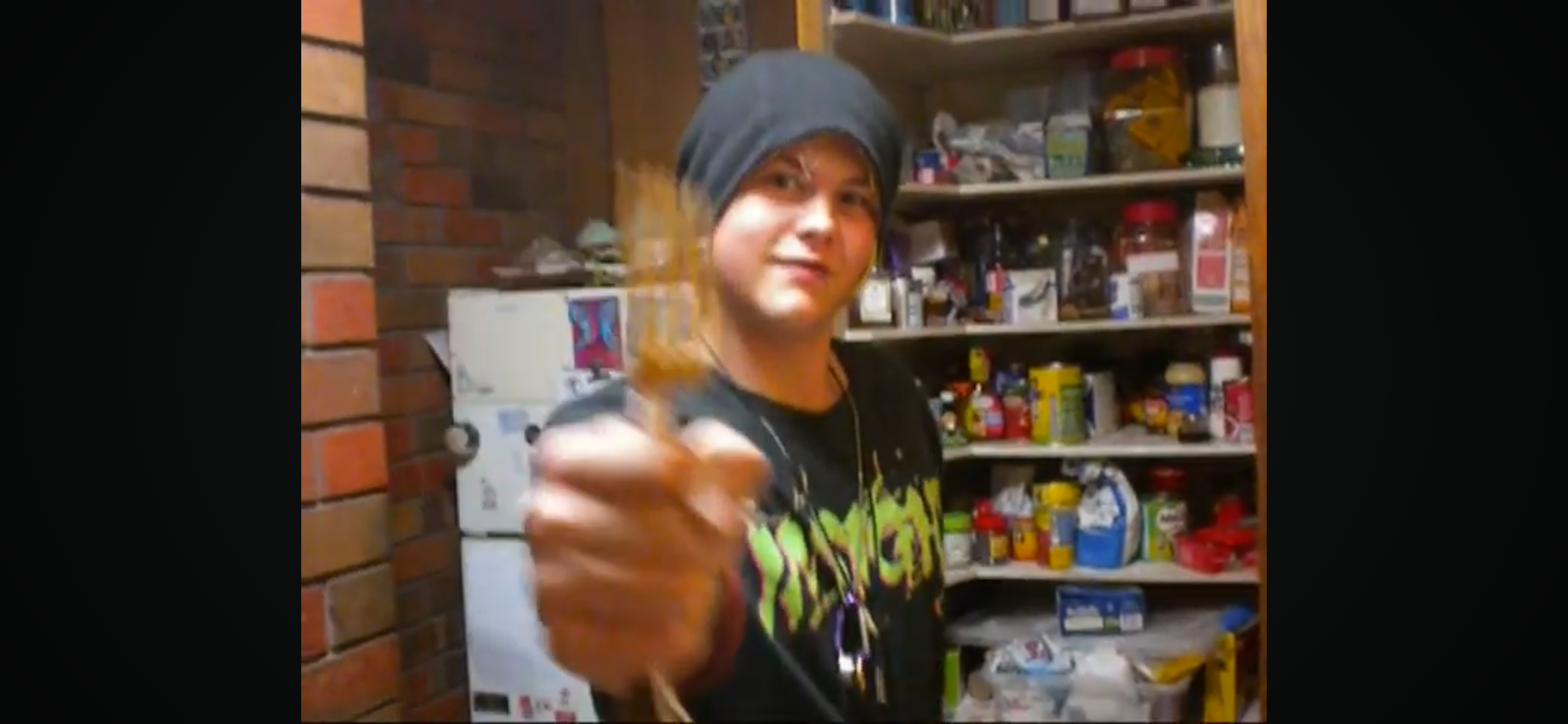 A person with a black hat and T-shirt pointing towards the camera in a pantry or storage room with shelves filled with various food and household items.