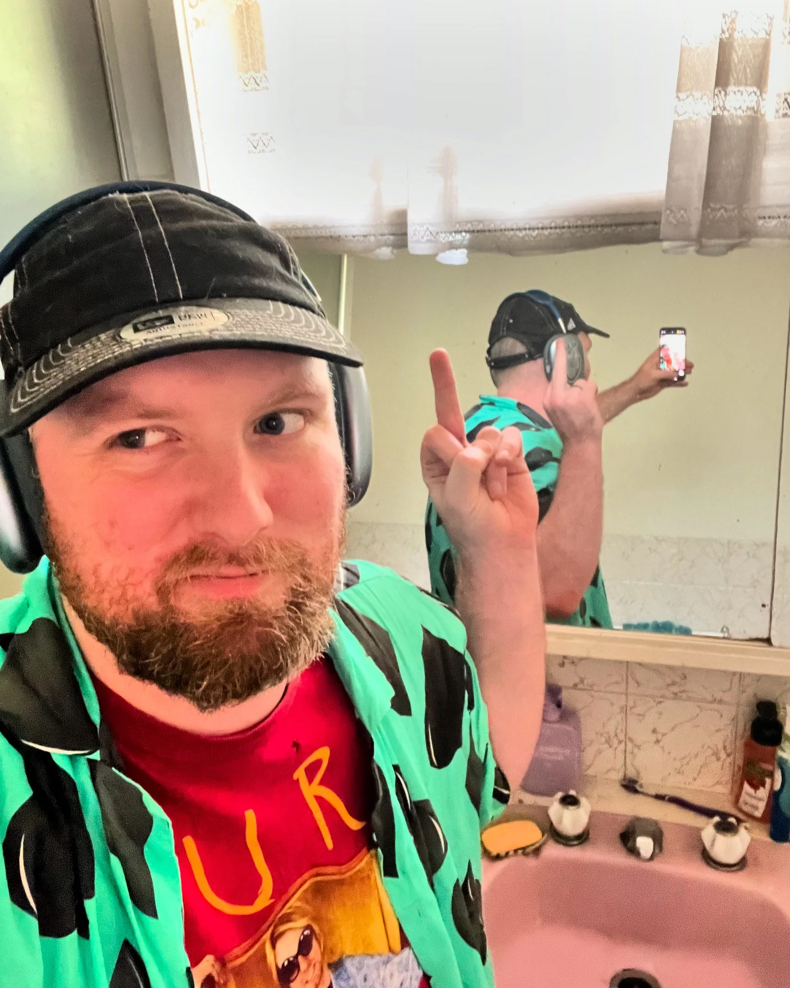 A man taking a selfie in a bathroom mirror. He's wearing a black cap, headphones, a red T-shirt, and a green patterned shirt. He is making a peace sign with his right hand.