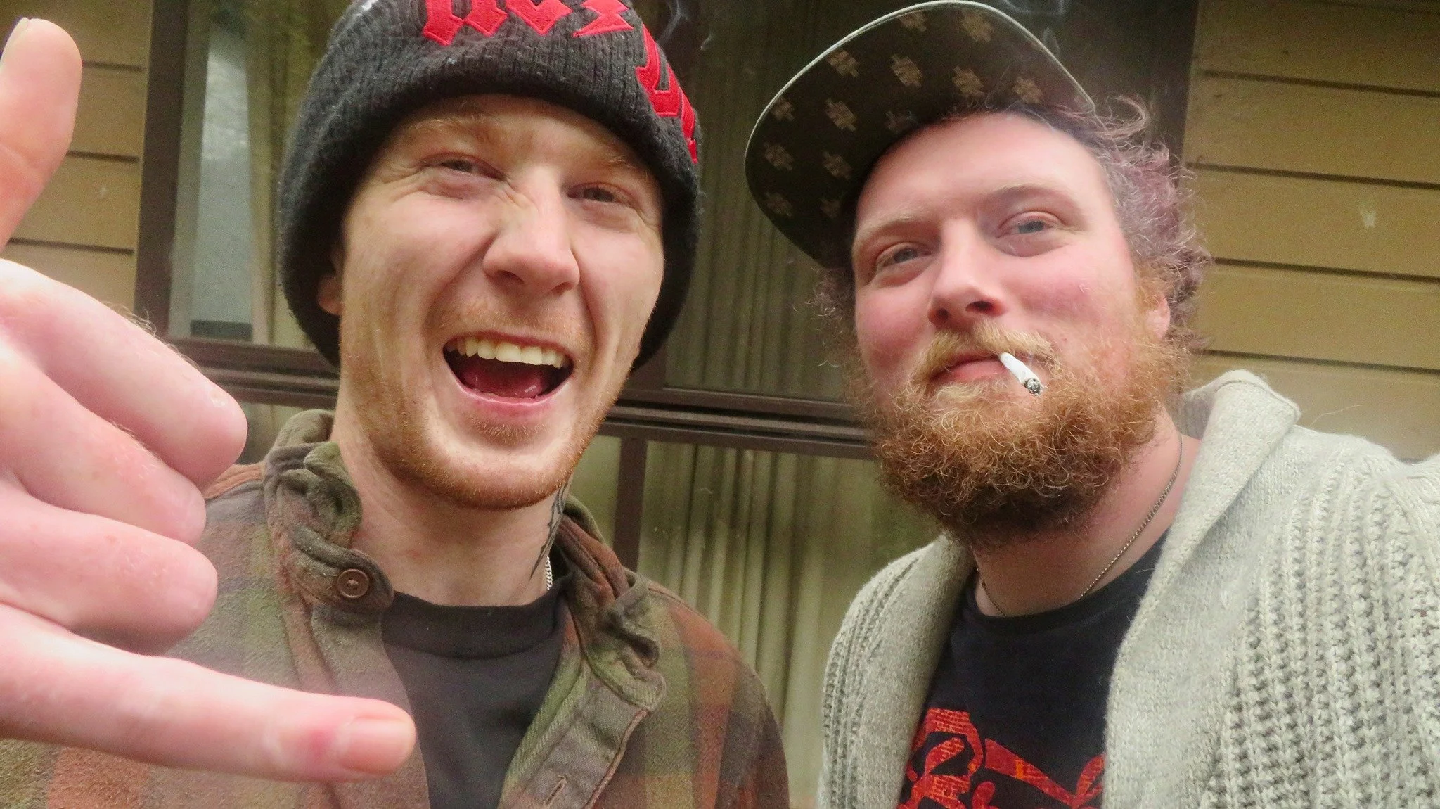Two men smiling for the camera; the man on the left is making a shaka sign with his hand, wearing a beanie, while the man on the right has a cigarette in his mouth, a beard, and a cap.