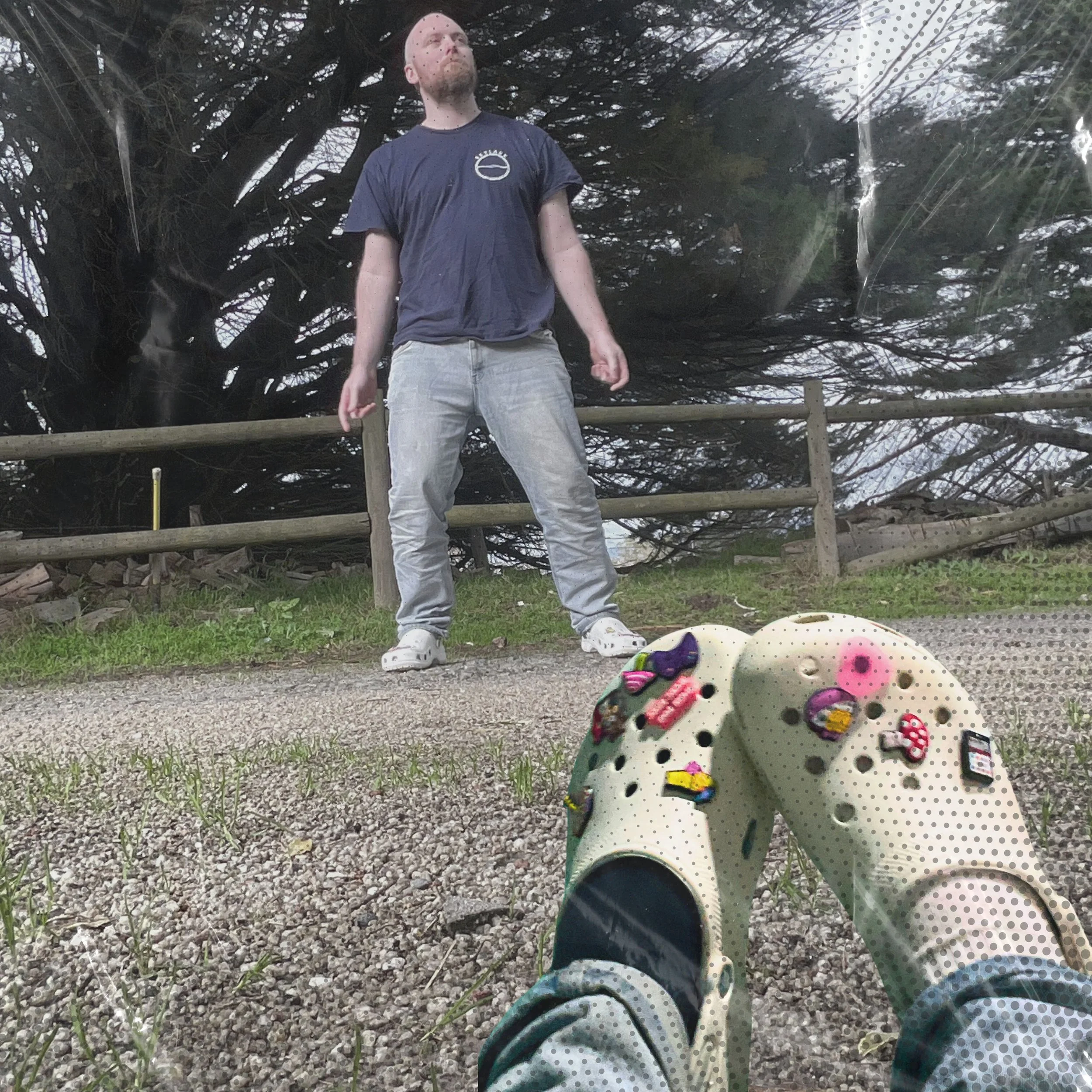 Person standing outdoors on a gravel surface with a wooden fence and large tree behind them, as seen from a ground-level perspective wearing patterned shoes with stickers.