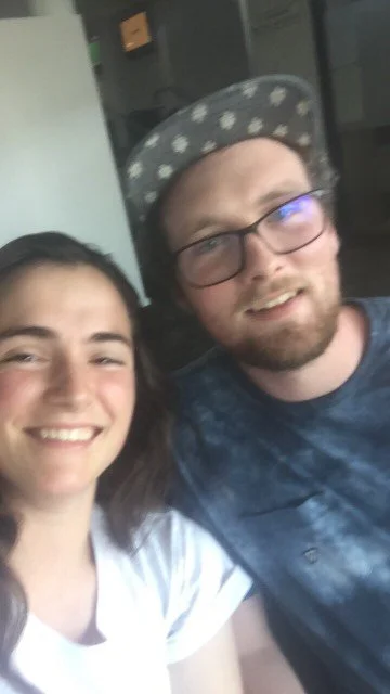 A young woman and a young man taking a selfie together indoors. The woman has long dark hair and is smiling, and the man has glasses, a beard, and is wearing a hat with a pattern.