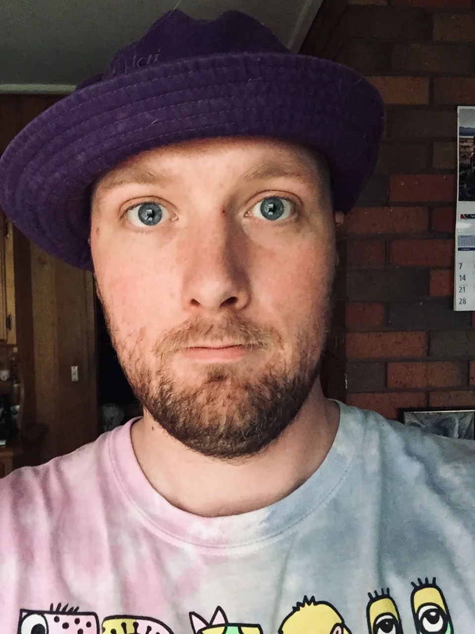A man with blue eyes, light skin, and a beard wearing a purple hat and a tie-dye t-shirt with cartoon characters, standing indoors in front of a brick wall and a calendar.