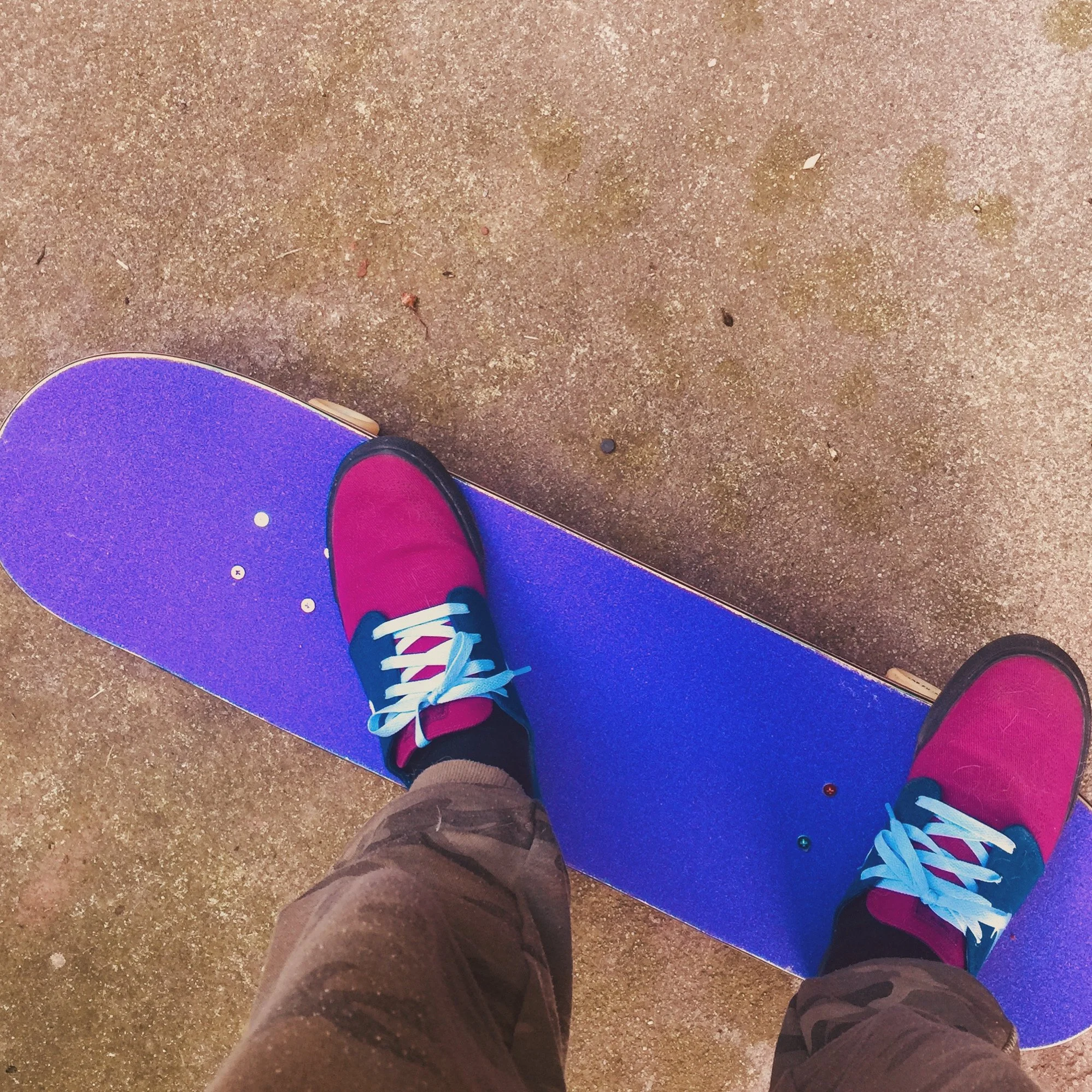 A person standing on a purple skateboard with pink shoes and camo pants on a concrete surface.