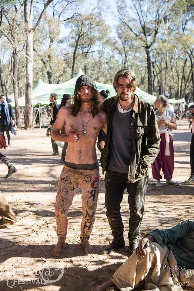 Two men standing outdoors at a festival, one shirtless with colorful tattoos on his legs and wearing a beanie, the other dressed in a jacket and jeans, with trees and tents in the background.