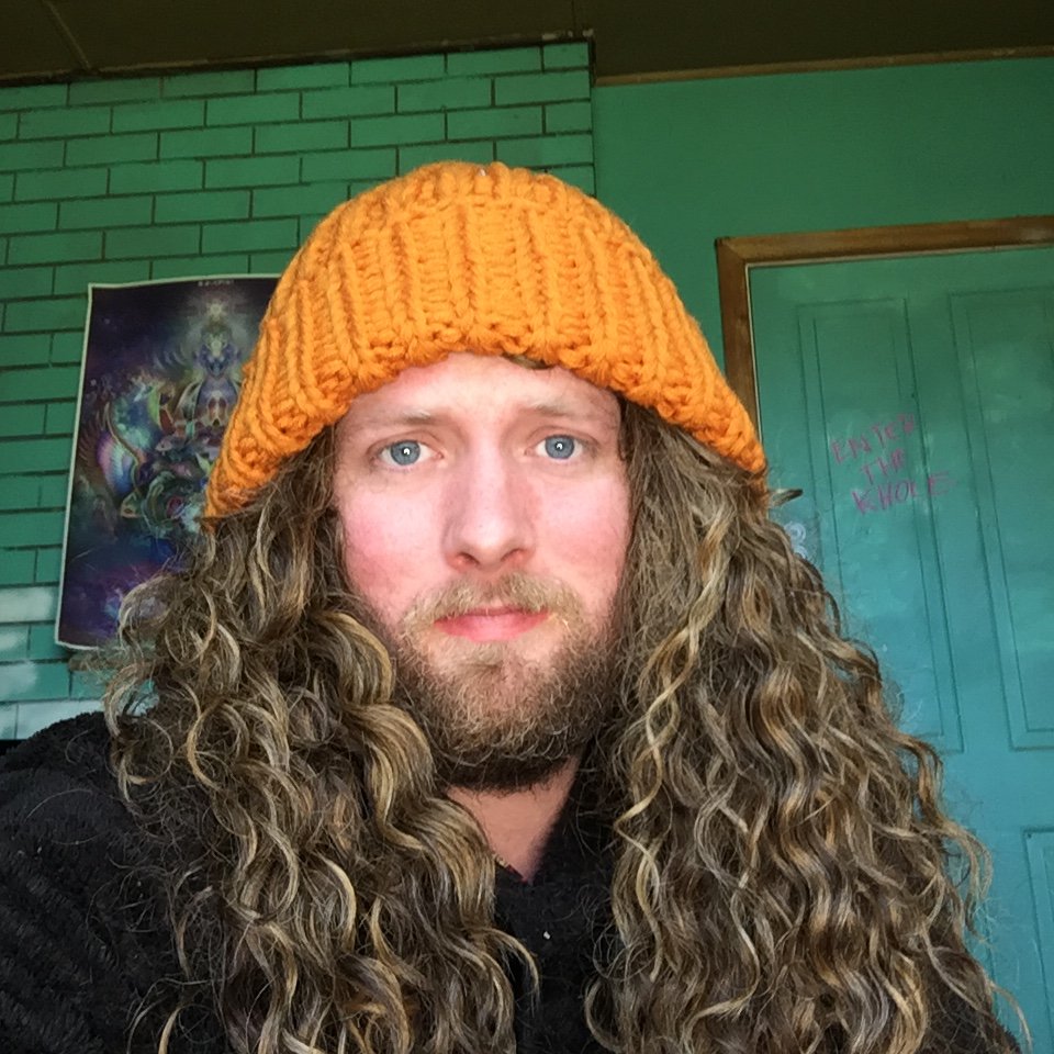 A man with long curly hair and a beard wearing an orange knitted beanie and black shirt, posing indoors in front of a teal green door and a wall with a colorful poster.