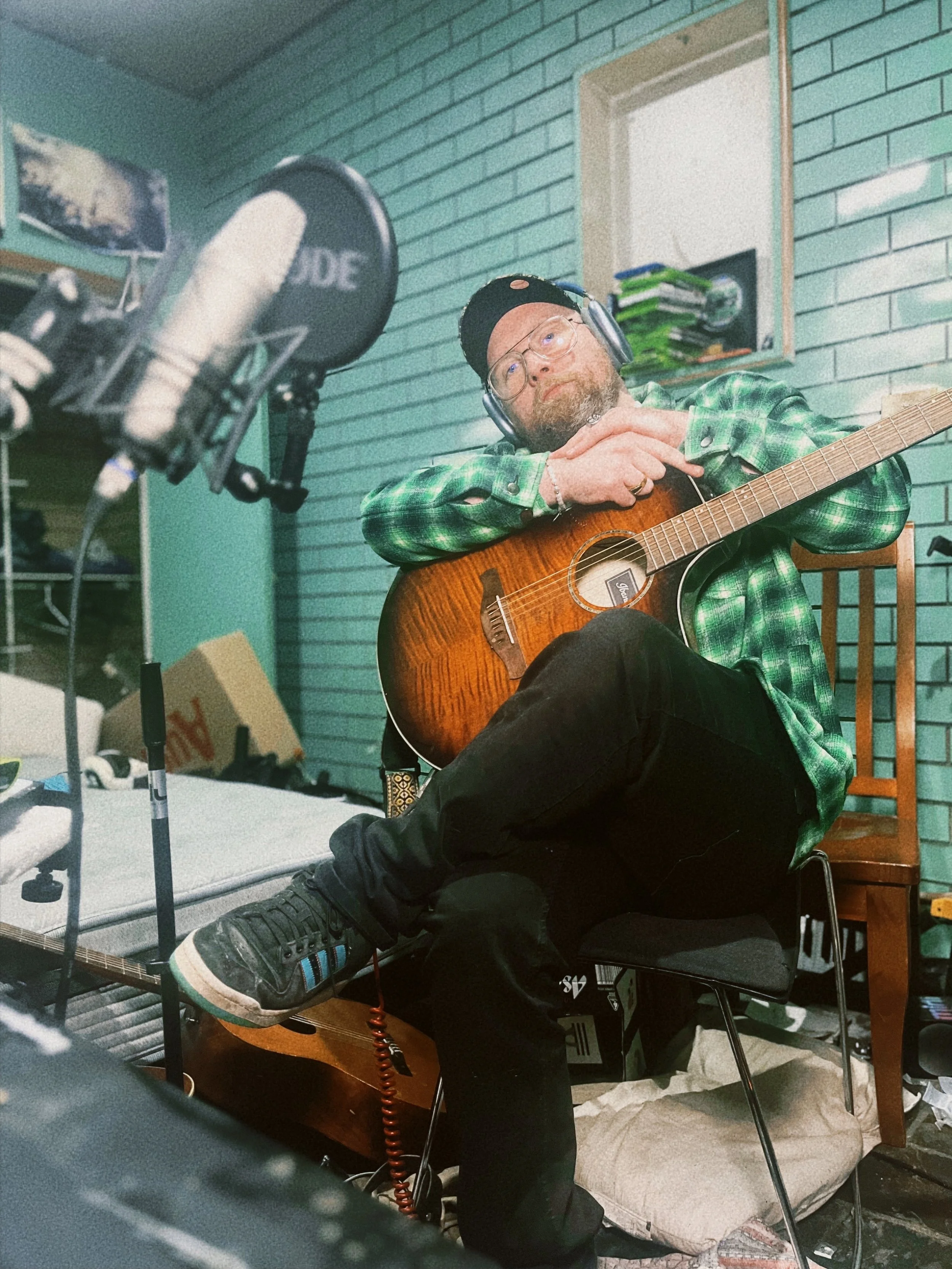 A man with glasses, a black cap, and a green checkered shirt playing an acoustic guitar in a room with brick walls. He is seated on a chair with his foot resting on an amplifier and is wearing headphones, surrounded by recording equipment.