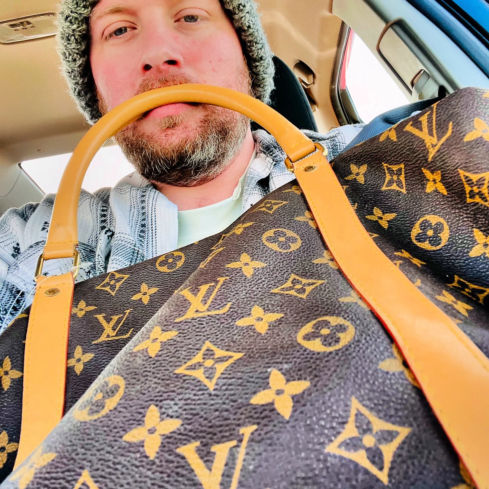 A man with a beard and wearing a knit hat, sitting inside a vehicle, holding a Louis Vuitton bag with a monogram pattern and tan handles.