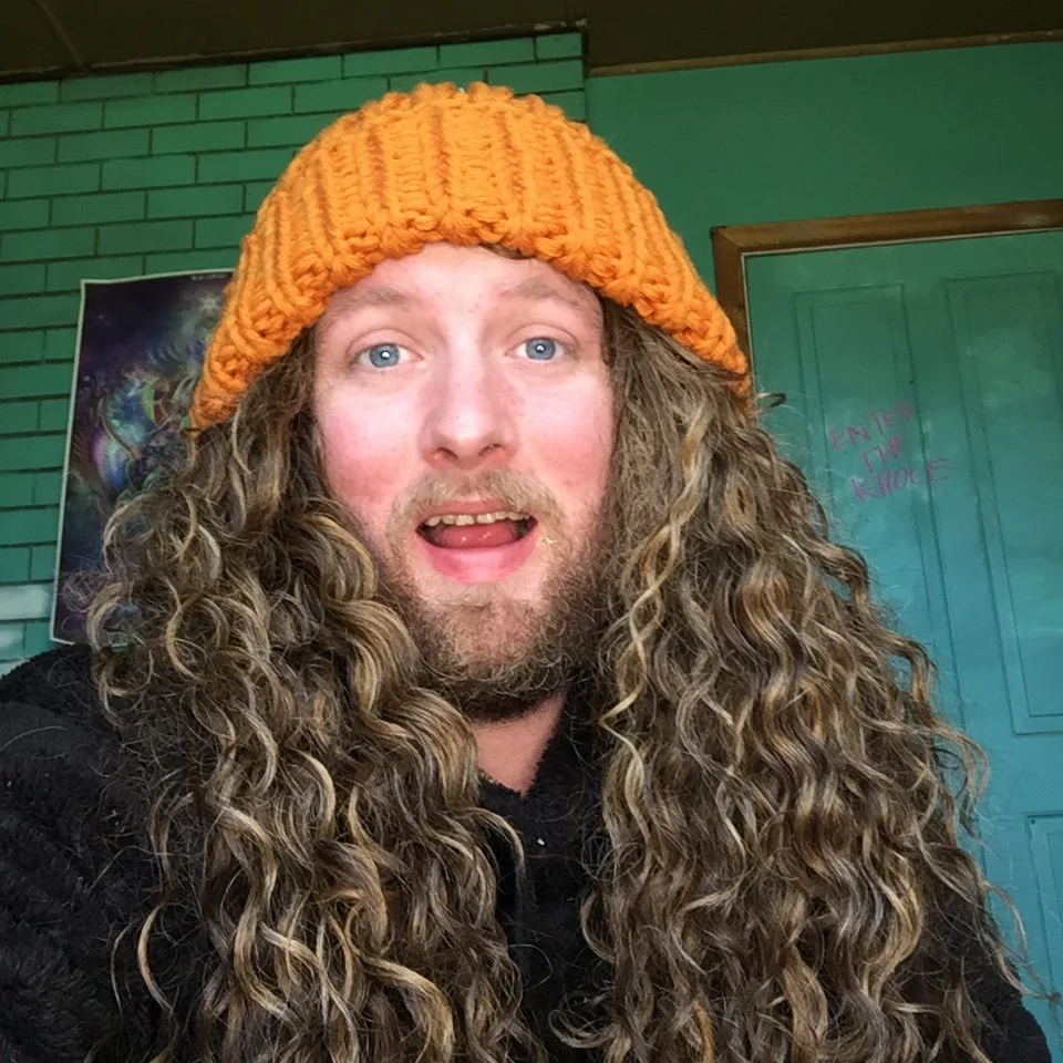 A man with long curly hair and a beard wearing an orange knitted beanie, smiling with his mouth open inside a colorful room.
