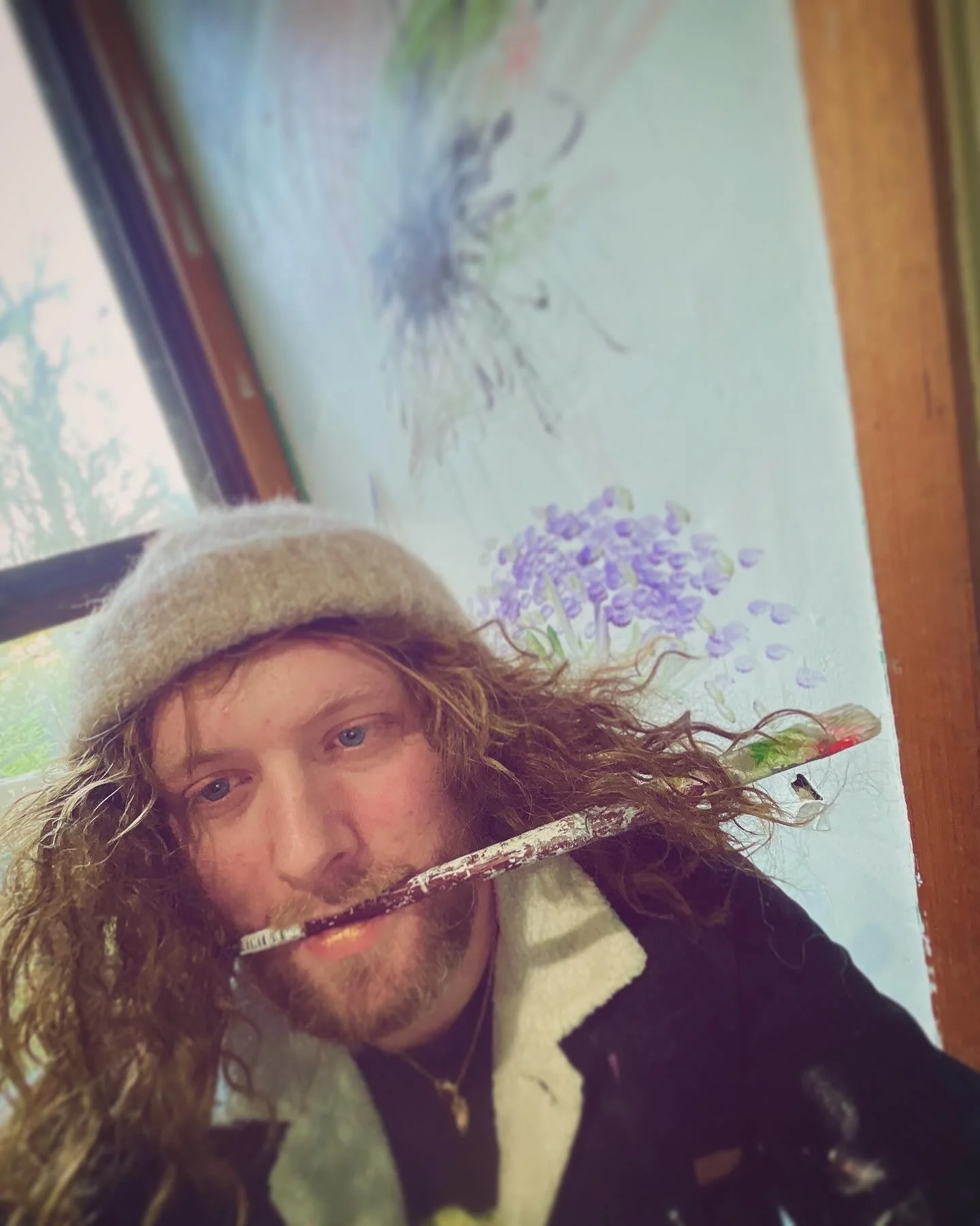 A person with long curly hair, wearing a beige beret and a black and white jacket, holding a paintbrush in his mouth, sitting in front of a window with a view of trees, and a watercolor painting of purple flowers on paper.