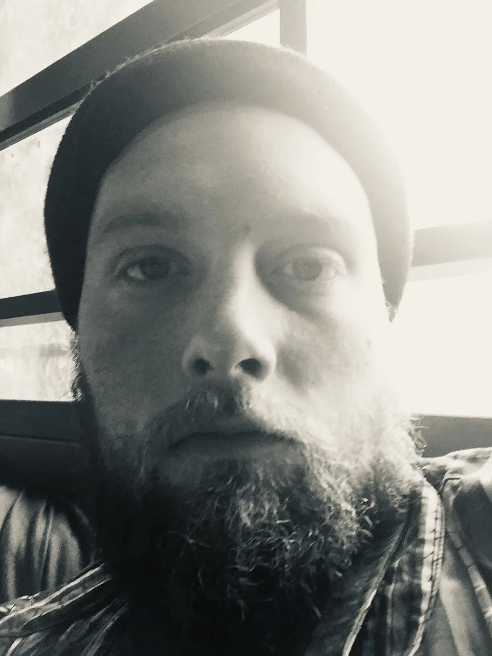 A close-up black-and-white photo of a man with a beard and mustache, wearing a cap, sitting near a window with sunlight shining through.