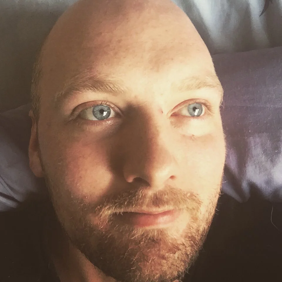 A close-up of a man with blue eyes, light skin, and a short beard, lying down on a pillow with sunlight illuminating his face.