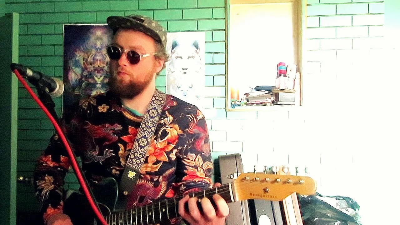 A man with a beard and sunglasses playing an electric guitar indoors, wearing a floral shirt and a patterned hat, with posters and tools visible in the background.