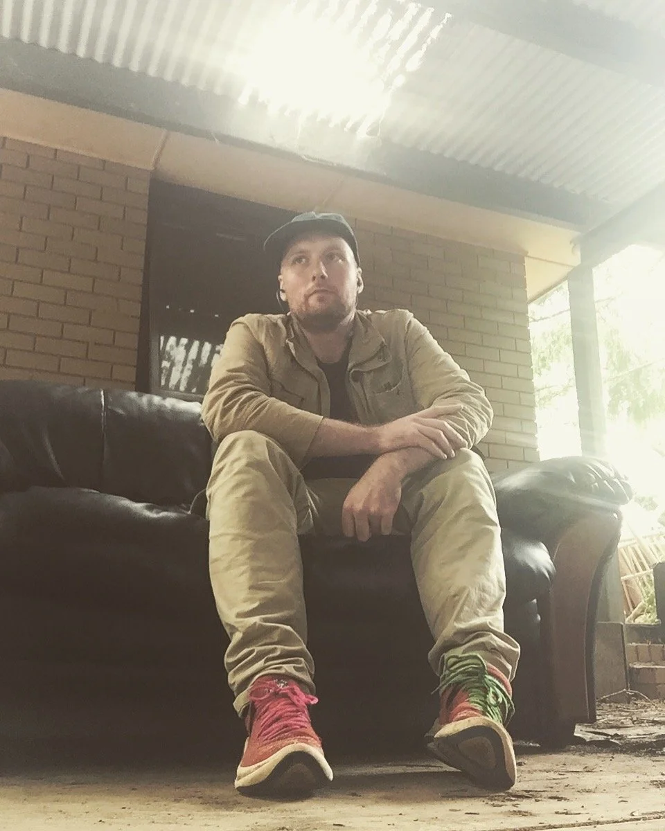 A man sitting on a black leather couch, wearing a beige jacket, beige pants, colorful sneakers, and a black cap, in a room with brick walls and a corrugated metal ceiling.