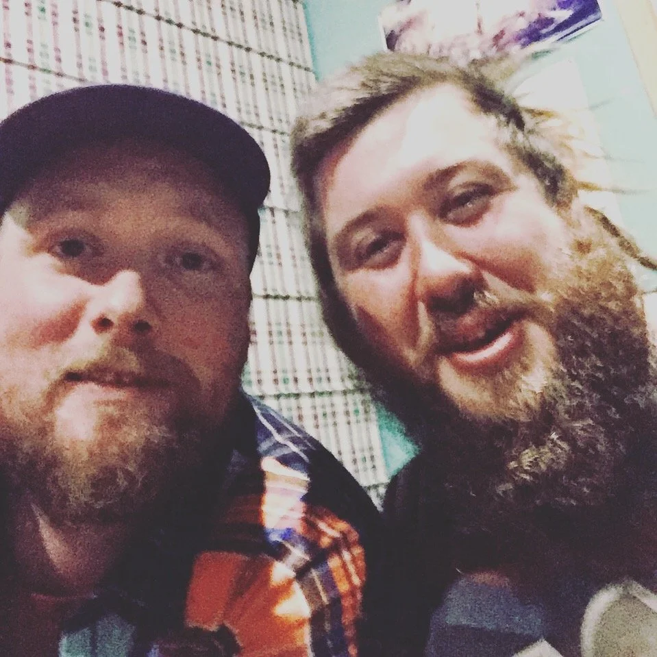 Two men with beards smiling and taking a selfie together indoors.