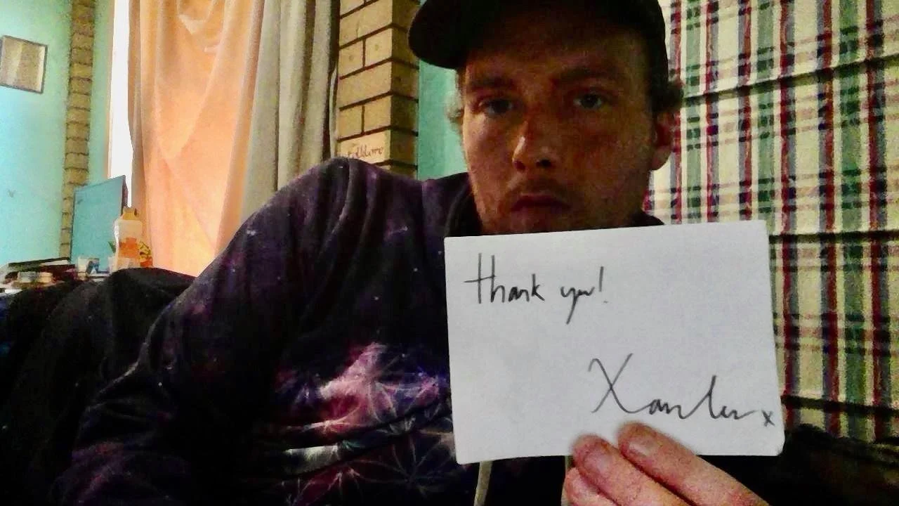 A man wearing a black cap holding a white card with handwritten text that says "thank you! XamLax" in a room with colorful curtains and a brick wall.