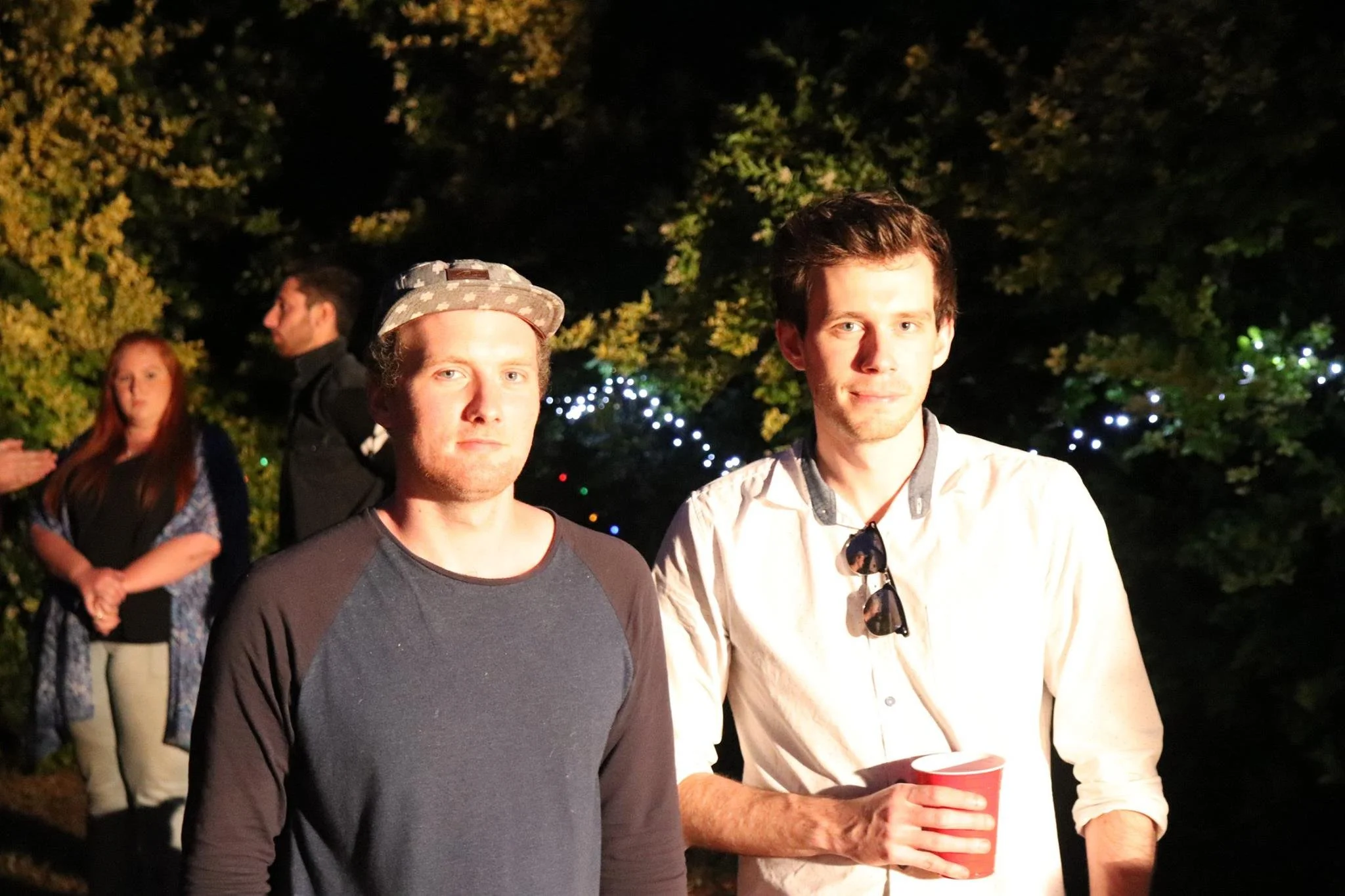 Two young men standing outdoors at night, one wearing a baseball cap and a dark long-sleeve shirt, the other in a white shirt with sunglasses hanging from the collar, holding a red cup. Two people are blurred in the background, with trees and string 