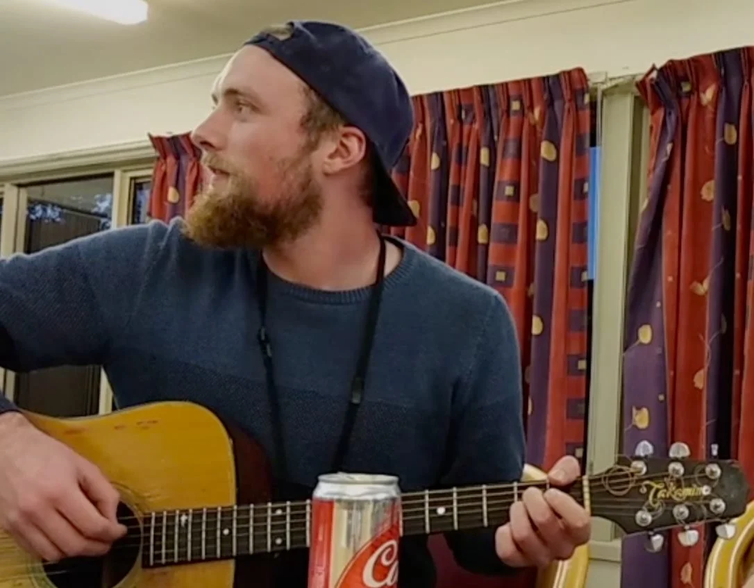 A man with a beard wearing a backward baseball cap playing an acoustic guitar indoors.