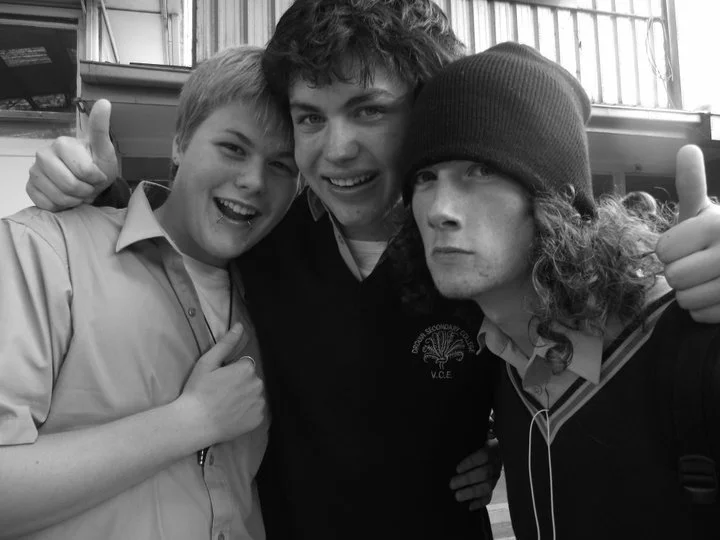 Three boys smiling and posing for a photo indoors, two giving thumbs up. The boy in the middle has curly hair and is wearing a dark shirt, the boy on the left has short hair and is wearing a light-colored shirt, and the boy on the right has long curl