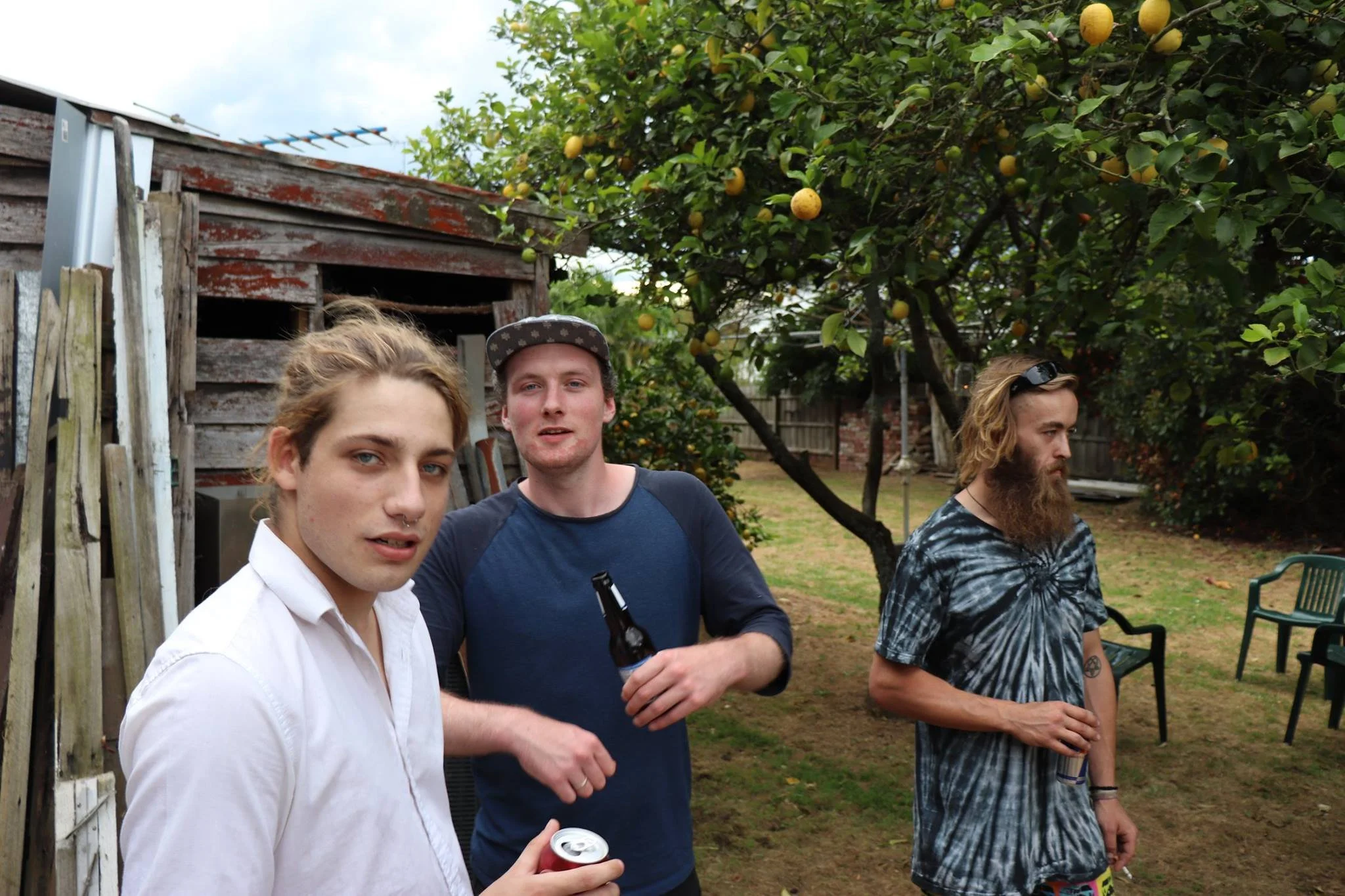 Three young men at an outdoor gathering in a backyard, near a lemon tree with ripe lemons. One is holding a drink and looking at the camera, another has a drink and faces forward, and the third has a drink and looks to the side. They are casually dre