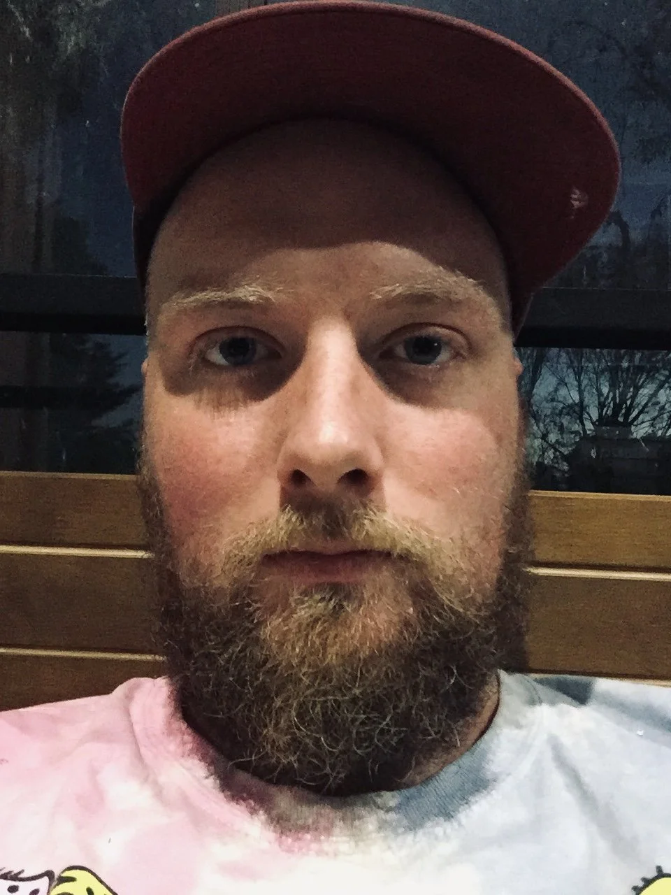 A close-up photo of a man with a beard, wearing a red cap, sitting in front of a window with trees outside and a wooden backrest behind him.