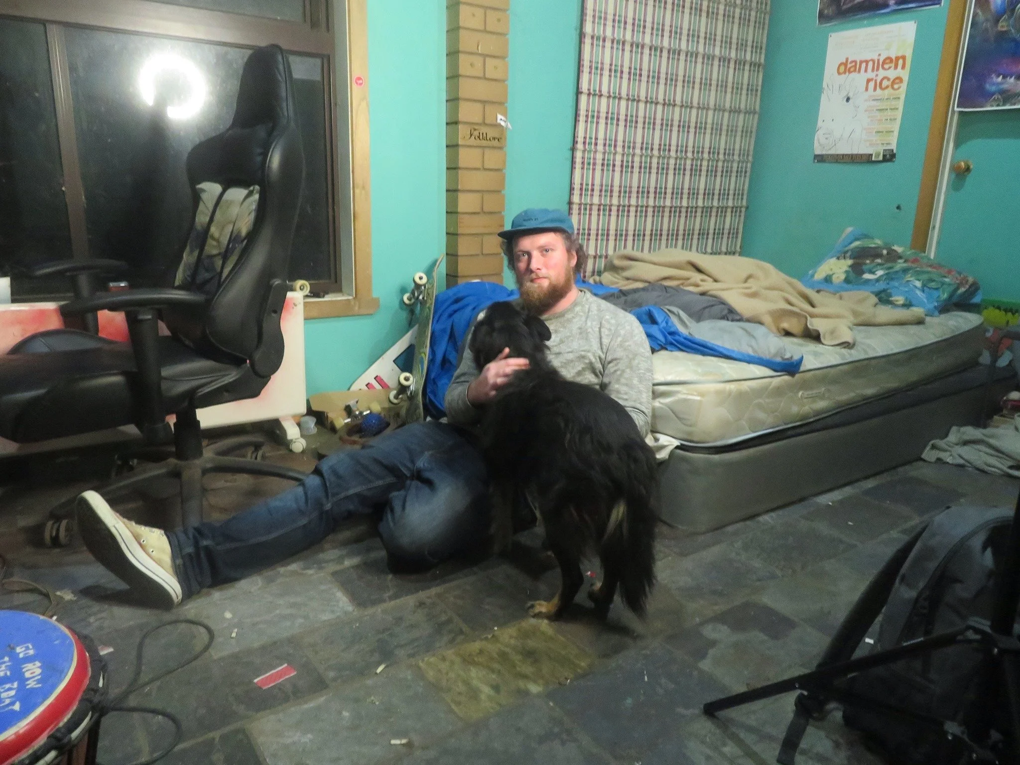 A man sitting on the floor with a black dog in a cluttered room, with a bed, skateboard, office chair, posters, and various items.