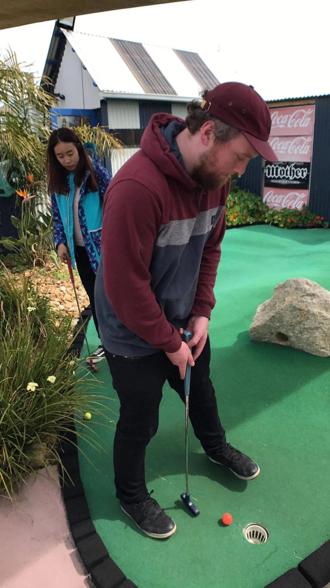 A man in a maroon and gray hoodie and cap playing miniature golf, preparing to putt a bright orange golf ball into the hole, with a woman in a blue jacket watching behind him.