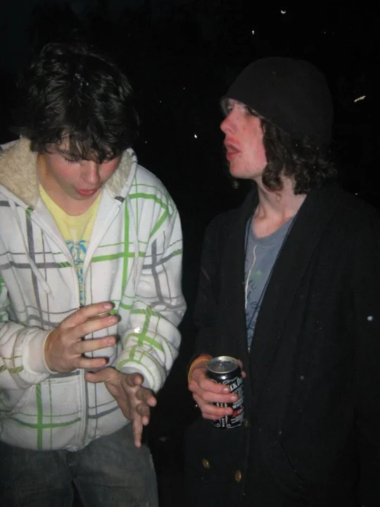 Two young men talking outdoors at night; one is wearing a white and green checkered hoodie and looking down at his hands, the other is wearing a black jacket and beanie, holding a soda can and engaging in conversation.