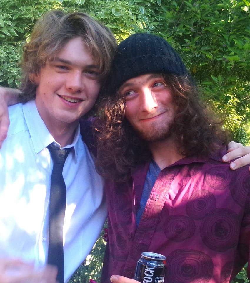 Two young men taking a selfie outdoors, one with light brown hair wearing a white shirt and black tie, the other with long curly hair, a black beanie, and a maroon patterned shirt, both smiling and posing closely surrounded by green foliage.