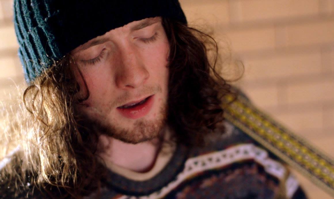 A young man with long, curly brown hair, wearing a black and blue striped knit cap, and a sweater with colorful patterns, appears to be playing an acoustic guitar.