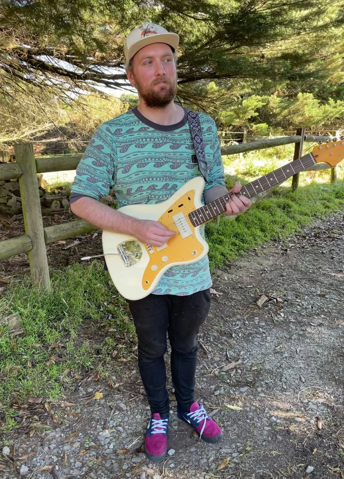 A man with a beard and mustache wearing a beige cap, a patterned blue T-shirt, black pants, and pink sneakers, standing outdoors on a dirt path beside a wooden fence, playing a white electric guitar with a yellow pickguard
