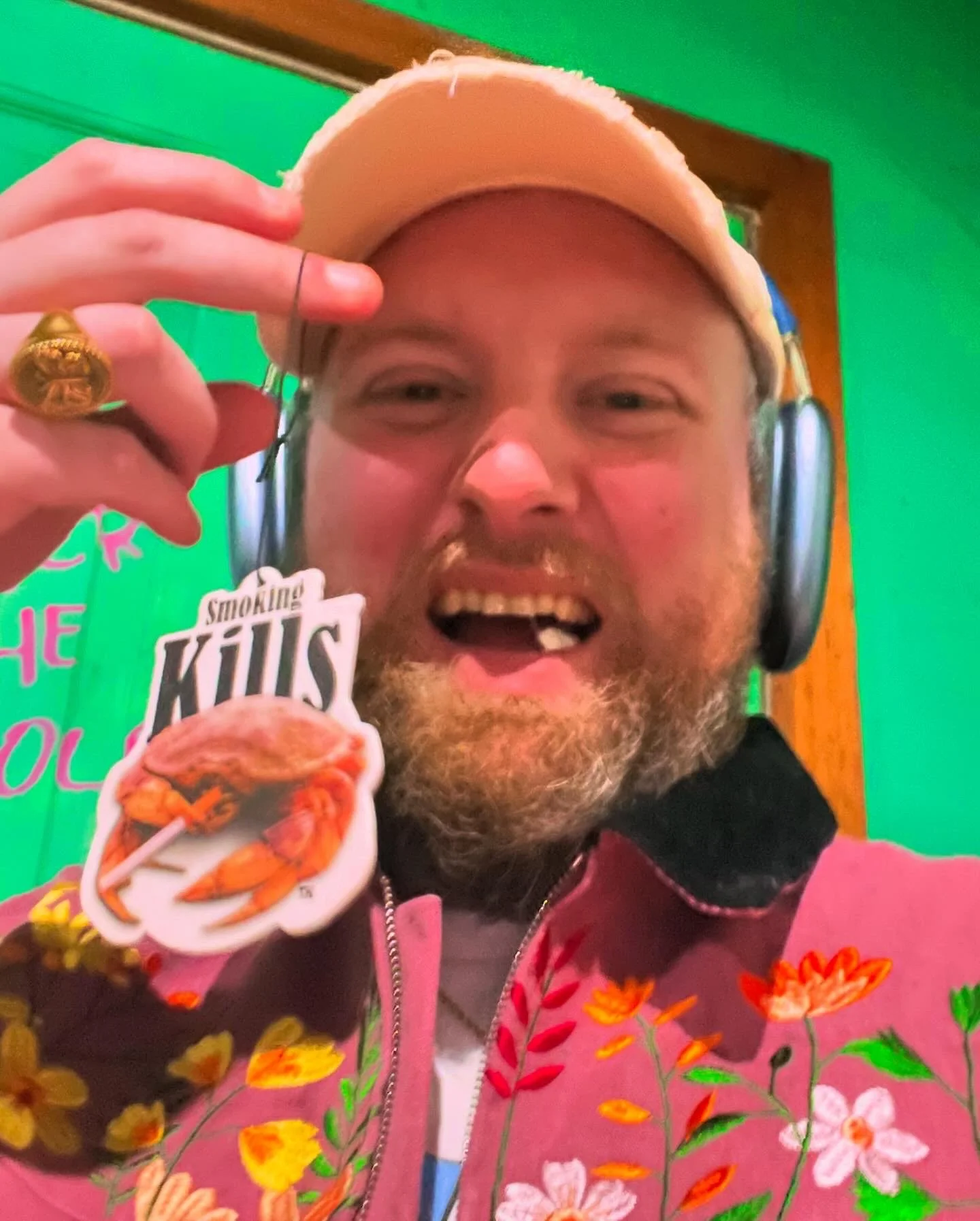 A man with a beard smiling, wearing a floral pink jacket, headphones, and a cap, holding a sticker of a crab with the text 'Smoking Kills' in front of a green background.