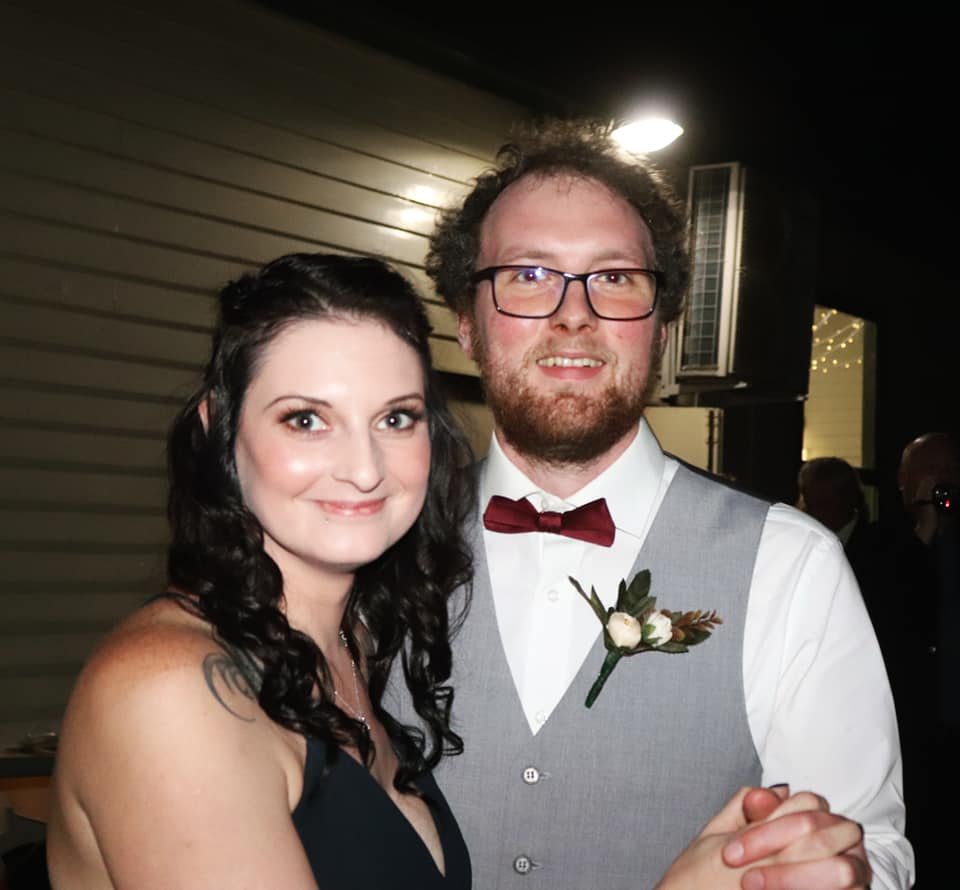 A smiling man and woman dancing together at a wedding reception. The man is wearing glasses, a white shirt, a gray vest, and a red bow tie with a boutonniere. The woman has dark, curly hair and is wearing a black dress.