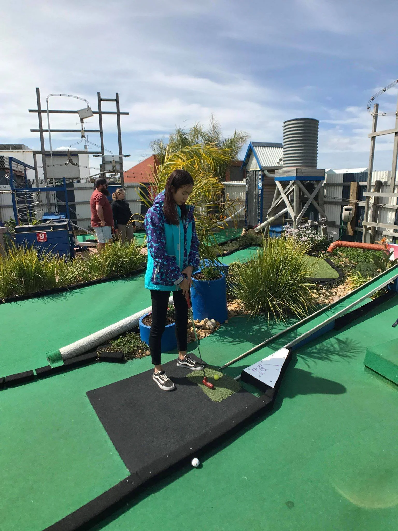 A young woman playing miniature golf on a green course, preparing to hit the ball with a club, with city buildings and a water tower in the background.