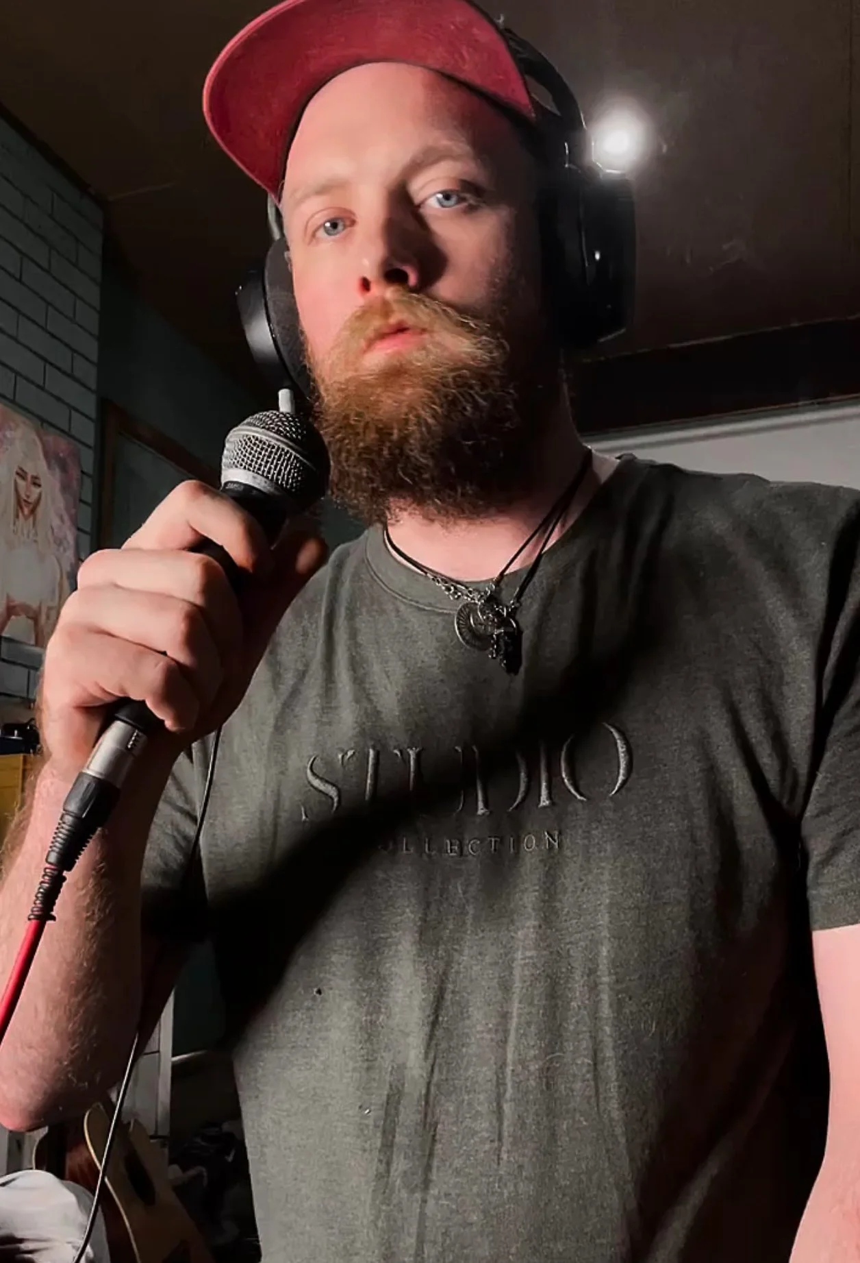 A man with a beard holding a microphone, wearing headphones, a red cap, and a dark T-shirt, standing indoors.