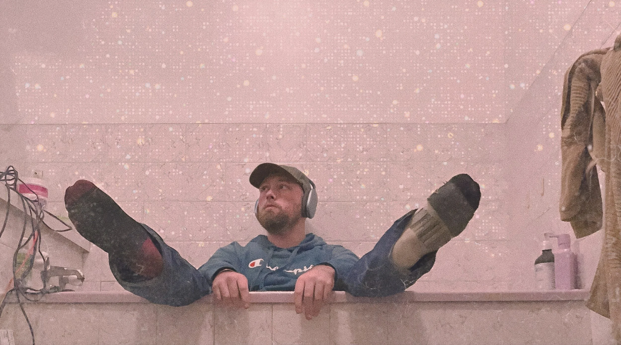 A man with a beard, wearing a cap, headphones, a blue Champion hoodie, and gloves, lying in a bathtub with legs stretched out and head resting on the edge, against a pink wall with sparkle-like patterns.