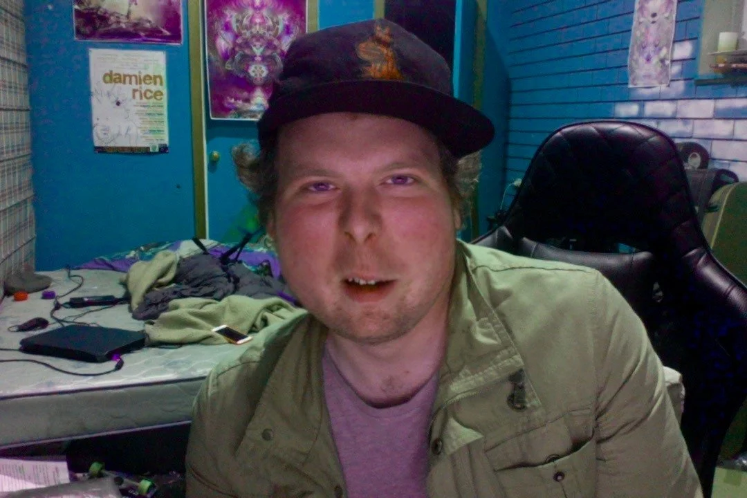 A man with curly hair wearing a baseball cap, tan jacket, and purple shirt sitting in a colorful room with posters and a bed in the background.