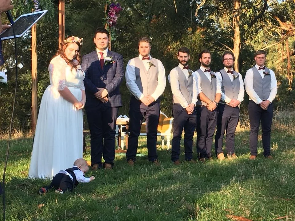 A wedding ceremony outdoors in a grassy area surrounded by trees. The bride wears a white dress with lace sleeves and a flower crown; she is pregnant. The groom and five groomsmen wear light vests and bow ties. A baby in a tux is crawling on the gras