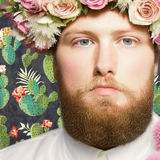 A man with a full beard and blue eyes wearing a floral crown made of pink and white roses, with a background of green prickly pear cactus plants.