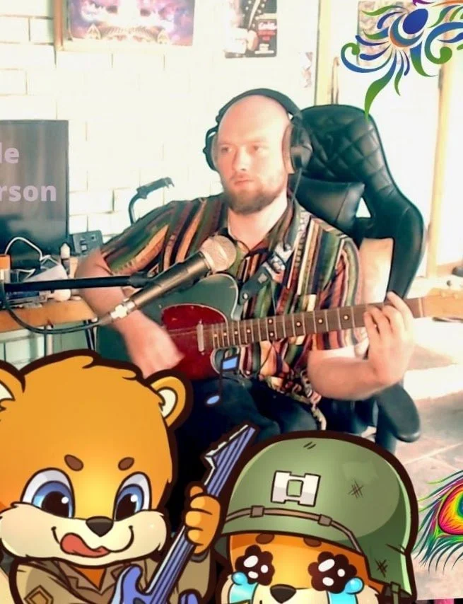 A man with a beard sitting in a black gaming chair, wearing headphones, playing an acoustic guitar and singing into a microphone. The background shows a white brick wall and some posters. Cartoon characters of a lion and a dog with a guitar and tears