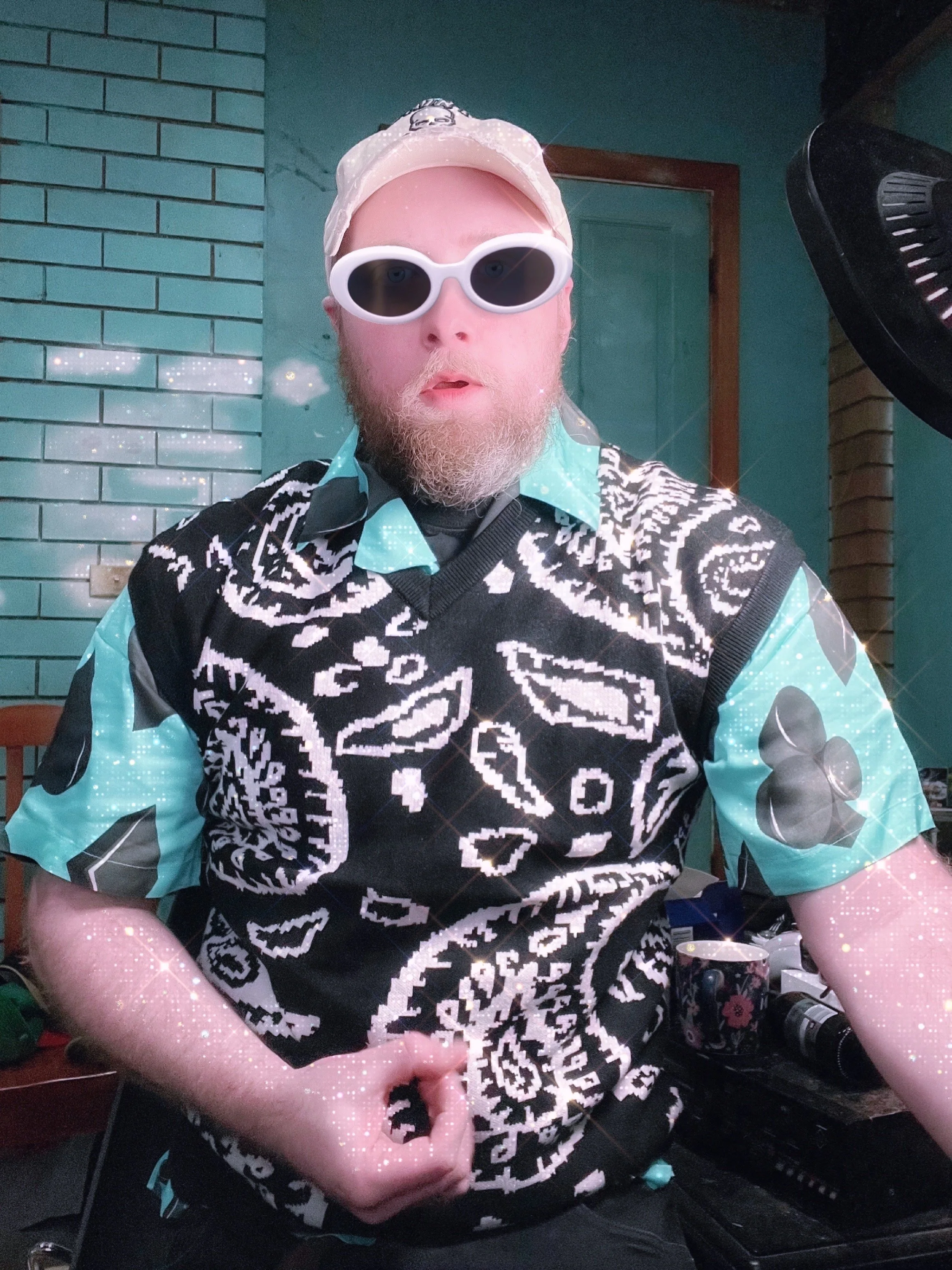 A man with a beard wearing white sunglasses, a beige cap, a black and white patterned sleeveless vest, and a turquoise shirt underneath. He is indoors, standing in front of a green brick wall, with a large black object and some electronic equipment v